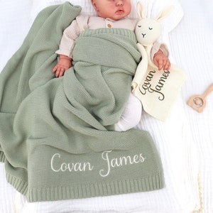 May include: A sleeping baby is wrapped in a sage green knitted blanket with the name "Covan James" embroidered in white. A matching security blanket and a stuffed bunny are nearby. A wooden rattle is also visible.