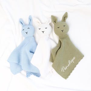 May include: Three knitted bunny security blankets in light blue, white, and olive green. Each has a bunny head with embroidered eyes and a blanket body. The olive green blanket has the name "Penelope" embroidered on it.