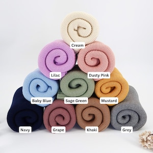 May include: A pyramid of rolled-up knitted blankets in various colors, including cream, lilac, dusty pink, baby blue, sage green, mustard, and grey. Each blanket is labeled with its color name. The blankets are arranged on a white background.