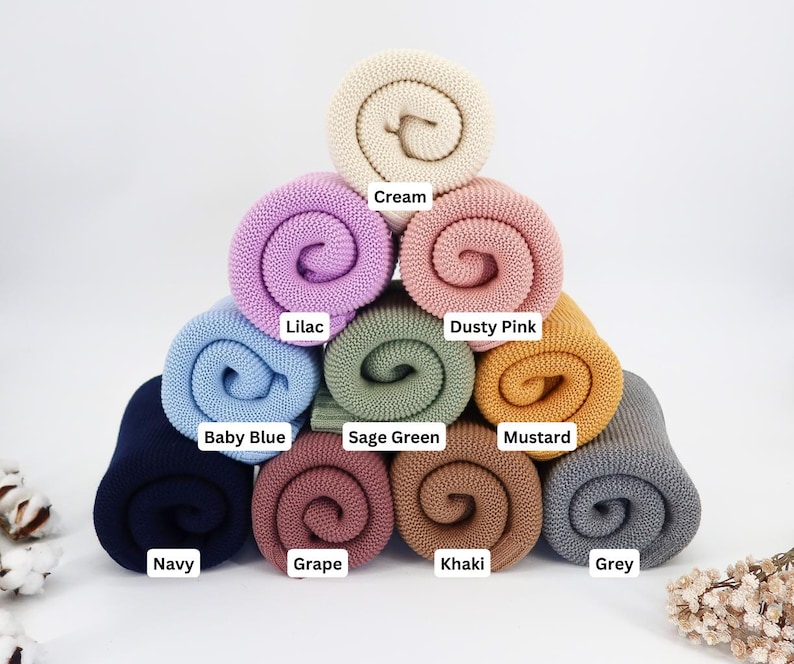 May include: A pyramid of rolled-up knitted blankets in various colors, including cream, lilac, dusty pink, baby blue, sage green, mustard, and grey. Each blanket is labeled with its color name. The blankets are arranged on a white background.
