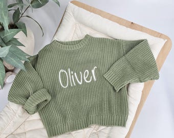 Może przedstawiać: Szałwiowy zielony sweter z dzianiny z imieniem "Oliver" wyhaftowanym białym pismem. Sweter jest prezentowany na kremowej, pikowanej powierzchni, z drewnianą ramą i rośliną doniczkową w tle. Sweter ma długie rękawy.