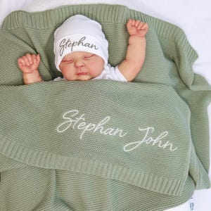 May include: A baby wrapped in a sage green knitted blanket and wearing a white beanie. The blanket and hat are embroidered with the name "Stephan" and "Stephan John" in white script. The baby is lying on a white surface.