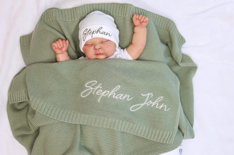 May include: A baby wrapped in a sage green knitted blanket and wearing a white beanie. The blanket and hat are embroidered with the name "Stephan" and "Stephan John" in white script. The baby is lying on a white surface.