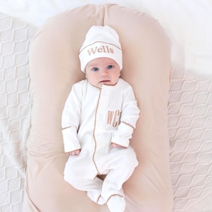 May include: A baby wearing a white onesie and matching hat, both with brown trim and embroidered details. The hat says "Wells" and the onesie has "WC" embroidered on the front. The baby is lying on a light pink pillow.