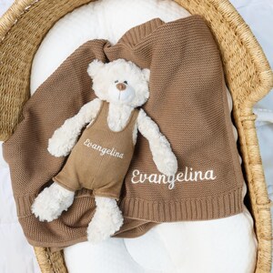 May include: A brown knitted baby blanket and a stuffed teddy bear with the name "Evangelina" embroidered on both. The bear wears brown overalls. The blanket and bear are in a woven basket.