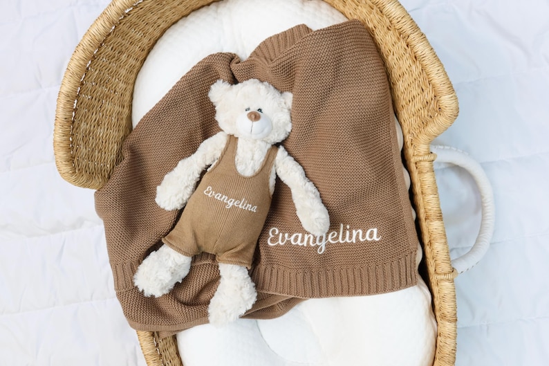 May include: A brown knitted baby blanket and a stuffed teddy bear with the name "Evangelina" embroidered on both. The bear wears brown overalls. The blanket and bear are in a woven basket.