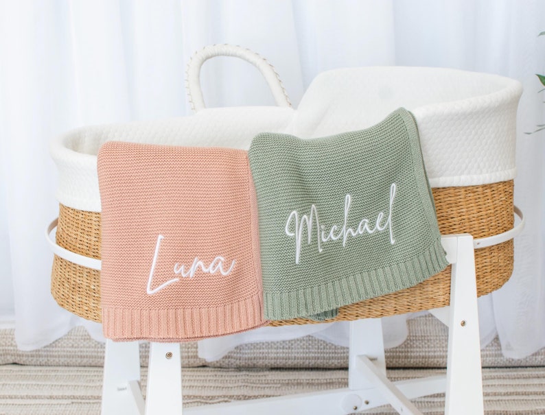 May include: Two personalized baby blankets, one in a soft pink and the other in a muted green, are draped over a white bassinet. The pink blanket has the name "Luna" embroidered in white, while the green blanket displays "Michael". The bassinet is made of woven wicker and white fabric.