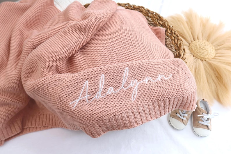 May include: A dusty rose knitted baby blanket with the name "Adalynn" embroidered in white script. The blanket is folded in a woven basket, with a pair of small tan baby shoes nearby. A decorative sunburst is in the background.