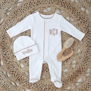 May include: White baby footed onesie and matching beanie with brown trim and embroidered initials and name. A wooden brush and comb set are also included. The items are displayed on a woven, circular rug.