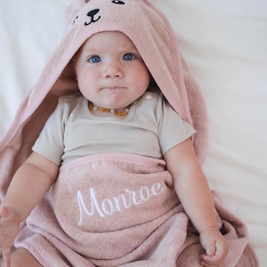 Peut inclure: Un b&eacute;b&eacute; envelopp&eacute; dans une serviette &agrave; capuche rose clair avec un motif de visage de lapin. Le nom "Monroe" est brod&eacute; en blanc sur la serviette. Le b&eacute;b&eacute; porte &eacute;galement un body de couleur claire.