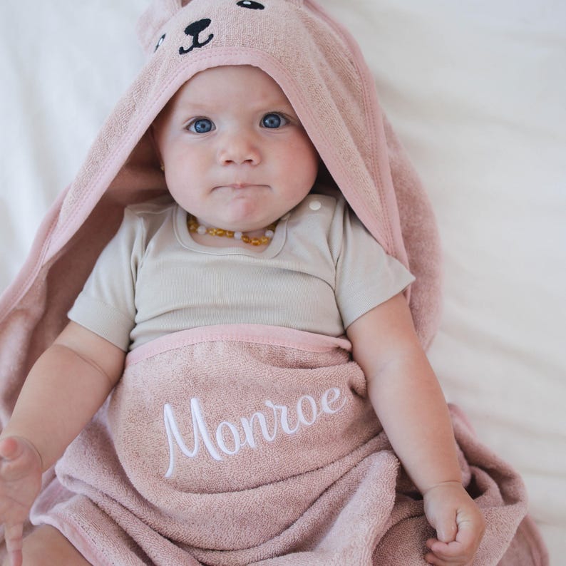 Peut inclure: Un b&eacute;b&eacute; envelopp&eacute; dans une serviette &agrave; capuche rose clair avec un motif de visage de lapin. Le nom "Monroe" est brod&eacute; en blanc sur la serviette. Le b&eacute;b&eacute; porte &eacute;galement un body de couleur claire.