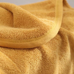 K&ouml;nnte beinhalten: Nahaufnahme eines gefalteten, goldgelben Handtuchs. Das Handtuch hat eine strukturierte, geschlungene Oberfl&auml;che und einen glatten, gen&auml;hten Rand. Der Stoff ist weich und wirkt saugf&auml;hig, geeignet f&uuml;r Bad oder Spa.