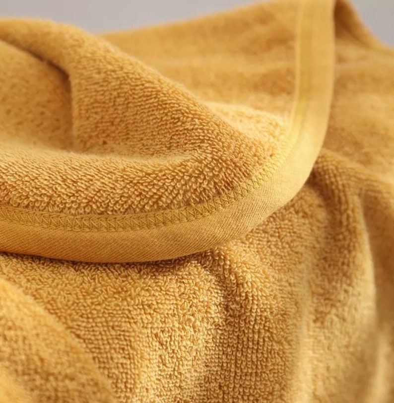 K&ouml;nnte beinhalten: Nahaufnahme eines gefalteten, goldgelben Handtuchs. Das Handtuch hat eine strukturierte, geschlungene Oberfl&auml;che und einen glatten, gen&auml;hten Rand. Der Stoff ist weich und wirkt saugf&auml;hig, geeignet f&uuml;r Bad oder Spa.