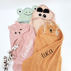 Peut inclure: Quatre serviettes &agrave; capuche pour b&eacute;b&eacute;s aux couleurs pastel, chacune avec un motif de visage d'animal. Une serviette rose avec un lapin et le nom "Caroline" brod&eacute;, une serviette panda beige, une serviette grenouille verte et une serviette lion beige avec le nom "Luca".
