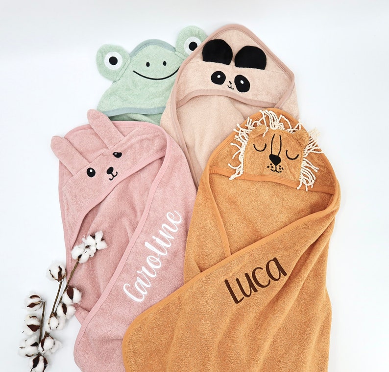 Peut inclure: Quatre serviettes &agrave; capuche pour b&eacute;b&eacute;s aux couleurs pastel, chacune avec un motif de visage d'animal. Une serviette rose avec un lapin et le nom "Caroline" brod&eacute;, une serviette panda beige, une serviette grenouille verte et une serviette lion beige avec le nom "Luca".