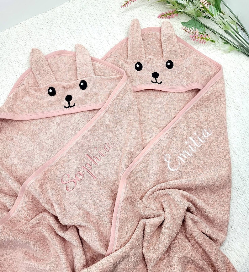 K&ouml;nnte beinhalten: Zwei rosa Kapuzenhandt&uuml;cher f&uuml;r Babys mit Hasenohren und gestickten Gesichtern. Die Handt&uuml;cher tragen die Namen "Sophia" und "Emilia" in Wei&szlig; gestickt. Die Handt&uuml;cher sind aus weichem Material und mit Rosa eingefasst.