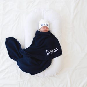 May include: A newborn baby wrapped in a navy blue knitted blanket and wearing a white beanie, both embroidered with the name "Brixton" in white. The baby is resting on a white pillow, set against a white background.