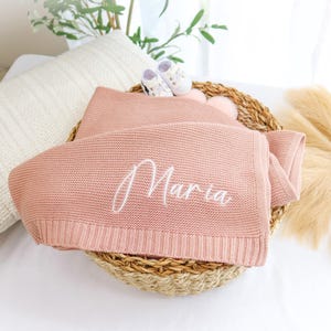 May include: A pink knitted baby blanket with the name "Maria" embroidered in white.