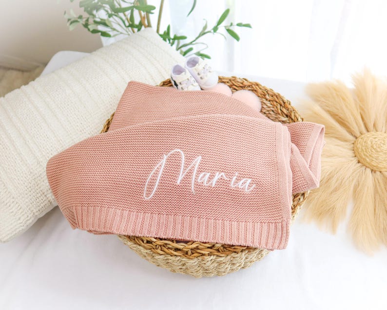 May include: A pink knitted baby blanket with the name "Maria" embroidered in white.