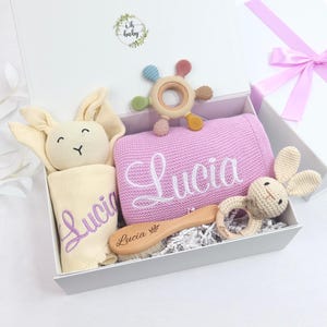 May include: A white gift box filled with baby items. Includes a pink blanket and a cream-colored bunny toy, both embroidered with the name "Lucia". Also features a wooden brush, a teether, and a crochet bunny rattle.