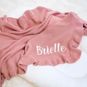 May include: A dusty rose-colored knitted baby blanket with a ruffled edge. The blanket is draped over a white pillow and features the name "Brielle" embroidered in white script. The blanket has a soft, textured appearance.