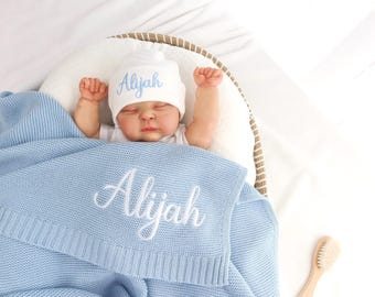 May include: A baby wrapped in a light blue knitted blanket and wearing a matching hat, both embroidered with the name "Aliyah" in white script. The baby is in a woven basket, with a wooden brush nearby.