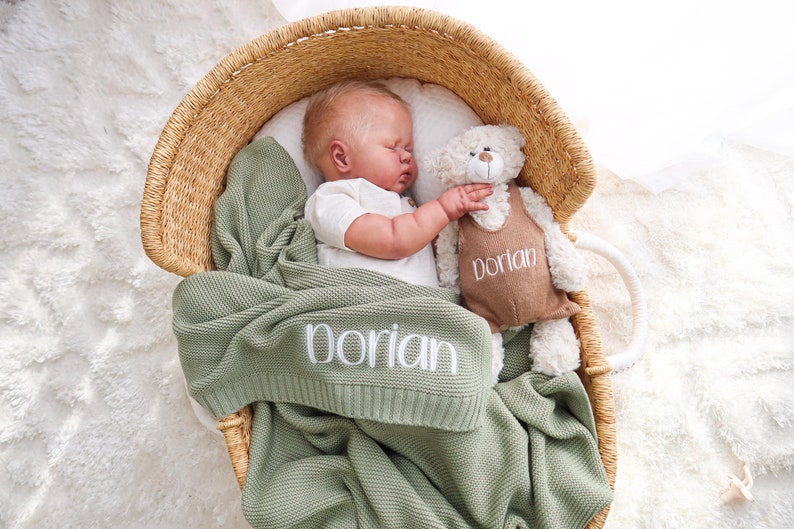 May include: A sleeping baby in a woven basket, nestled with a sage green blanket embroidered with "Dorian". A plush teddy bear wearing a brown outfit with "Dorian" is beside the baby. The basket is on a white, fluffy surface.