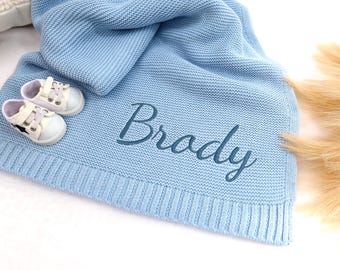 May include: A light blue knitted baby blanket with the name "Brady" embroidered on it. Two pairs of white baby shoes with black star accents are placed on the blanket. The blanket is folded and has a ribbed border.