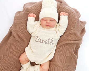 May include: A newborn baby wearing a cream-colored knitted outfit, including a hat with a pom-pom, a long-sleeved onesie, and booties. The name "Zareth" is embroidered on the onesie. The baby is lying on a brown knitted blanket.