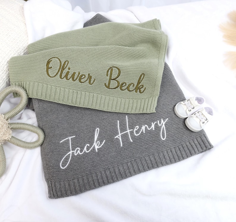 May include: Two personalized knitted baby blankets. One is sage green with the name "Oliver Beck" embroidered in gold thread. The other is gray with the name "Jack Henry" embroidered in white thread.