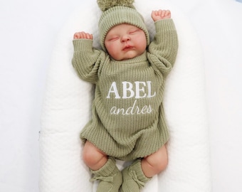 May include: A baby wearing a sage green knit outfit, including a hat with a pom-pom, a long-sleeved romper, and booties. The romper is embroidered with the name "ABEL Andres" in white. The baby is lying on a white, cushioned surface.