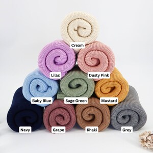 May include: A collection of rolled knit blankets in various colors, including cream, lilac, dusty pink, baby blue, sage green, mustard, navy, grape, khaki, and grey. Each blanket is labeled with its color name.