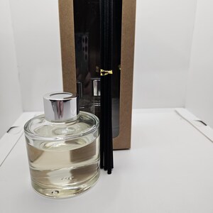 May include: A clear glass reed diffuser bottle with a silver cap filled with light-colored liquid, accompanied by a bundle of black reed sticks tied with a gold band, positioned in front of a tall rectangular brown cardboard box with a window.