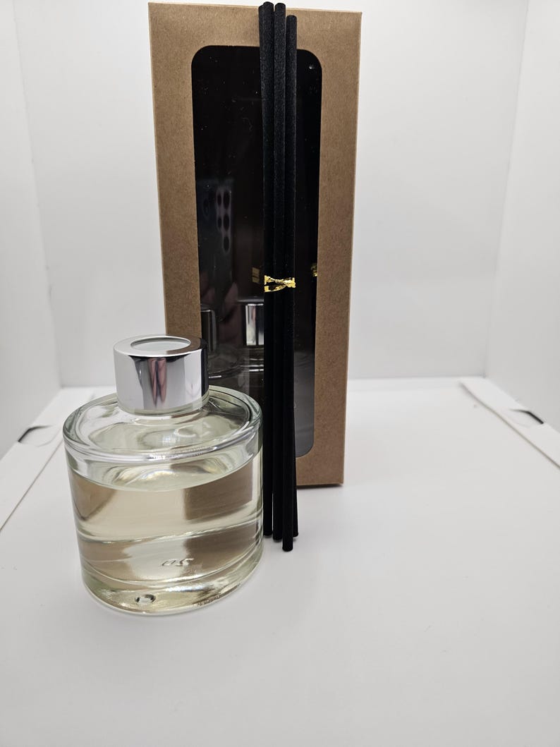 May include: A clear glass reed diffuser bottle with a silver cap filled with light-colored liquid, accompanied by a bundle of black reed sticks tied with a gold band, positioned in front of a tall rectangular brown cardboard box with a window.