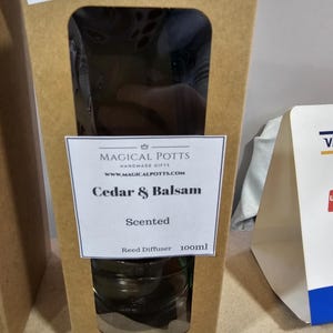 May include: A brown cardboard box containing a 100ml reed diffuser. The label reads Magical Potts Handmade Gifts, www.magicalpotts.com, Cedar & Balsam, Scented, Reed Diffuser 100ml. The box has a rectangular cutout window showing the glass bottle inside.