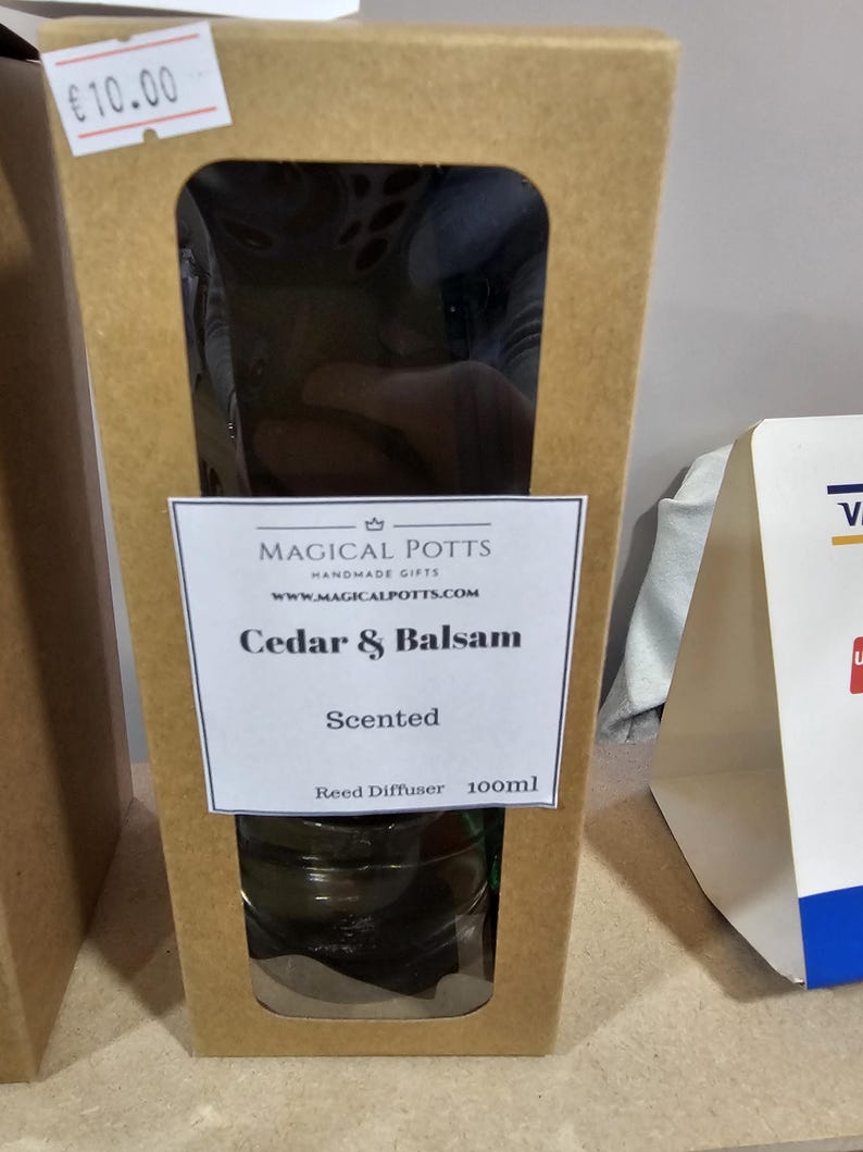 May include: A brown cardboard box containing a 100ml reed diffuser. The label reads Magical Potts Handmade Gifts, www.magicalpotts.com, Cedar & Balsam, Scented, Reed Diffuser 100ml. The box has a rectangular cutout window showing the glass bottle inside.