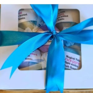 Puede incluir: Una caja de regalo blanca atada con una cinta azul brillante. La caja contiene varias bombas de baño, visibles a través de una ventana transparente. Las bombas de baño son de varios colores y tienen etiquetas que dicen "bombas perfumadas" y "Magical Pots Ever Glow Apple scented Bath Bombs."