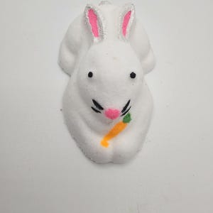 Könnte beinhalten: Ein weißes, hasenförmiges Marshmallow-Konfekt mit rosa gefütterten Ohren, schwarzen Augen, einer rosa Nase und einer kleinen orangefarbenen Karotte. Die Süßigkeit zum Thema Ostern liegt auf einer weißen Oberfläche.