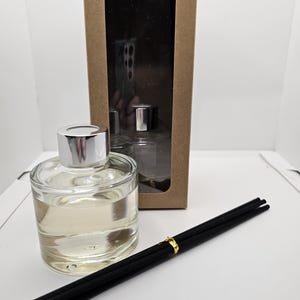 May include: A clear glass reed diffuser bottle with a silver cap, filled with a light-colored liquid. Beside it lies a bundle of black reed sticks secured with a gold band. Behind the bottle stands a tall, rectangular brown cardboard box.