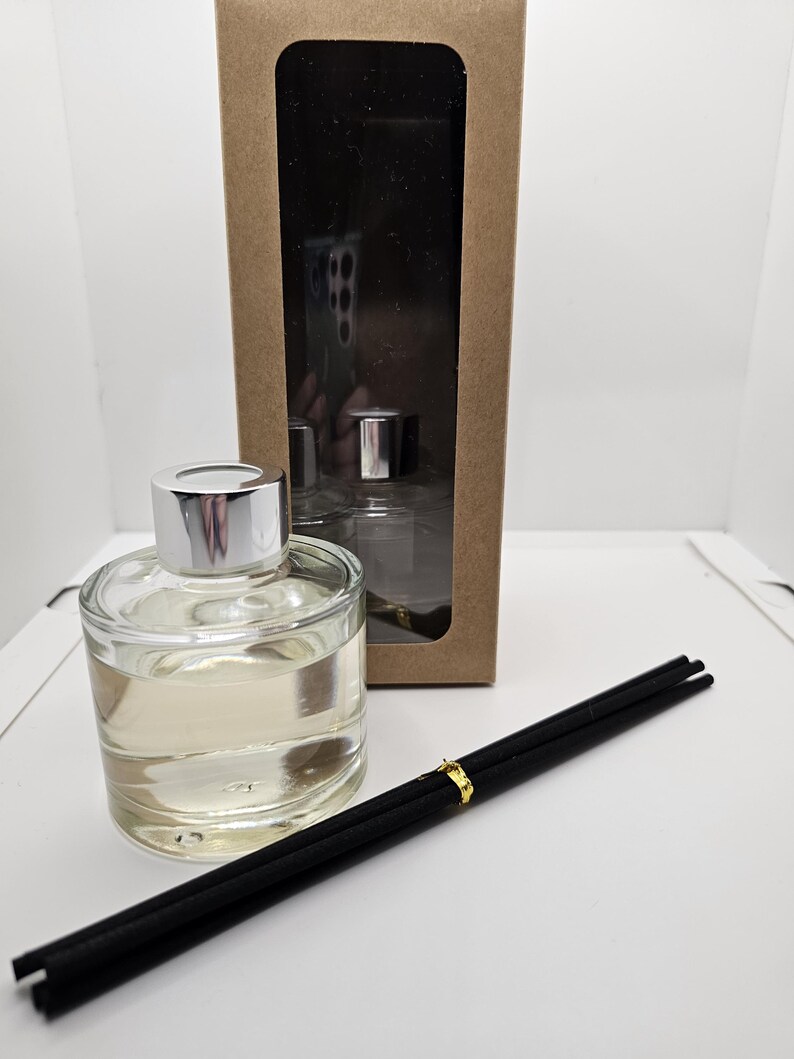 May include: A clear glass reed diffuser bottle with a silver cap, filled with a light-colored liquid. Beside it lies a bundle of black reed sticks secured with a gold band. Behind the bottle stands a tall, rectangular brown cardboard box.