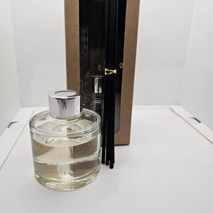 May include: A clear glass reed diffuser bottle with a silver cap filled with a light-colored liquid, accompanied by a set of five black reed sticks tied with a gold band, all presented in front of a tall rectangular brown cardboard box.
