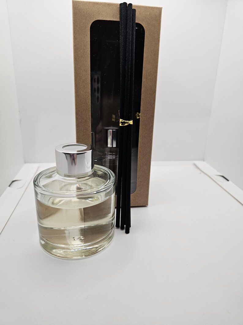 May include: A clear glass reed diffuser bottle with a silver cap filled with a light-colored liquid, accompanied by a set of five black reed sticks tied with a gold band, all presented in front of a tall rectangular brown cardboard box.