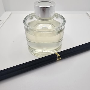 May include: A clear glass reed diffuser bottle with a silver cap, filled with a pale liquid. Three black reed sticks are bundled together with a small gold-colored band, positioned diagonally in front of the glass container.