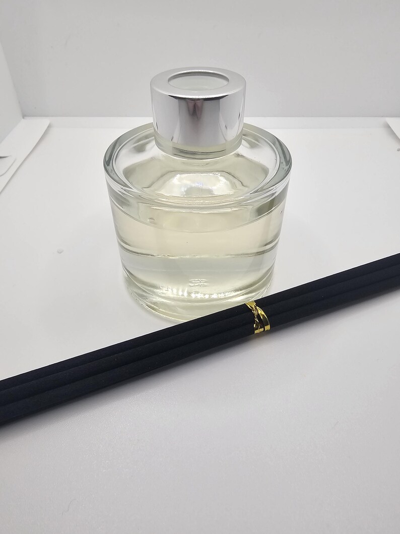 May include: A clear glass reed diffuser bottle with a silver cap, filled with a pale liquid. Three black reed sticks are bundled together with a small gold-colored band, positioned diagonally in front of the glass container.