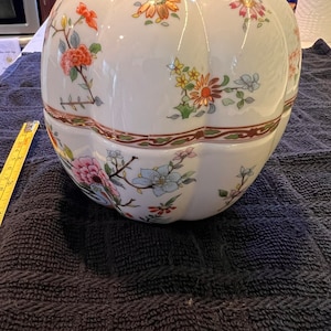May include: A white ceramic pumpkin-shaped container with a lid. The container is decorated with floral designs in shades of pink, blue, yellow, and green. The lid has a gold trim.