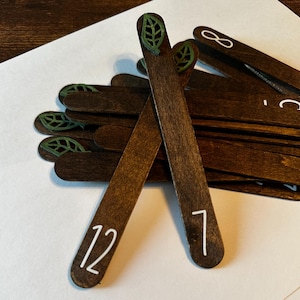 May include: A set of wooden sticks with green leaf designs and white numbers, including the numbers 7, 8, 12, and others.