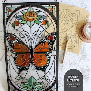 May include: A stained glass pattern featuring a butterfly with orange and black wings, a rose with orange petals, and green leaves. The pattern is framed in black metal and is ready to be used for a stained glass project.