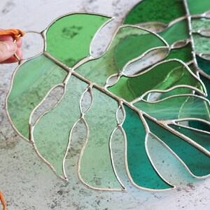 Peut inclure: Deux feuilles de monstera en verre teint&eacute; dans des tons de vert, avec un cordon en cuir marron attach&eacute; au sommet d'une feuille.
