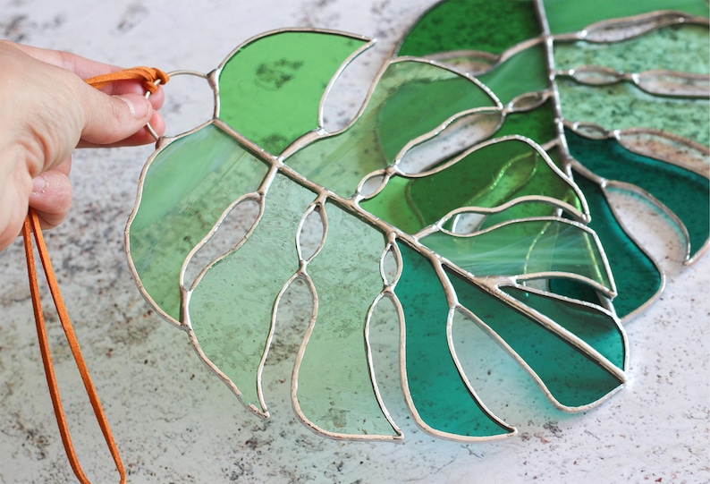 Peut inclure: Deux feuilles de monstera en verre teint&eacute; dans des tons de vert, avec un cordon en cuir marron attach&eacute; au sommet d'une feuille.