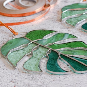Peut inclure: Deux feuilles de monstera en verre teint&eacute; vert avec des accents en feuille d'argent. Les feuilles sont suspendues &agrave; un cordon en cuir brun.